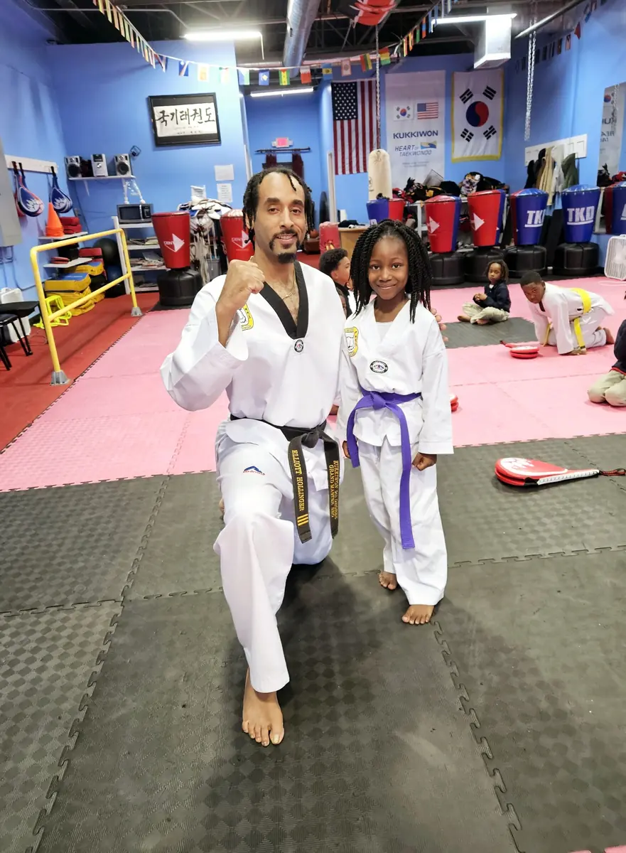 A black-belt instructor kneeling beside a young purple-belt student at Master Pius Martial Arts